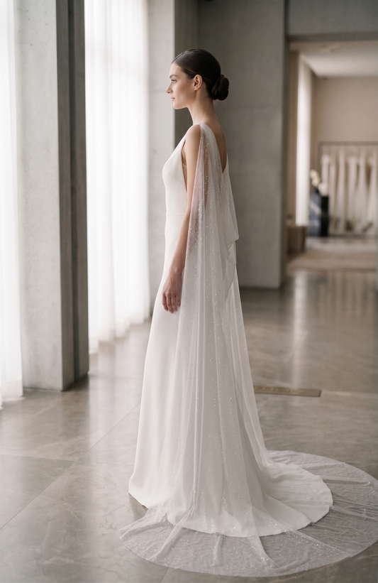 Woman in a white wedding dress standing in a softly lit room with large windows.