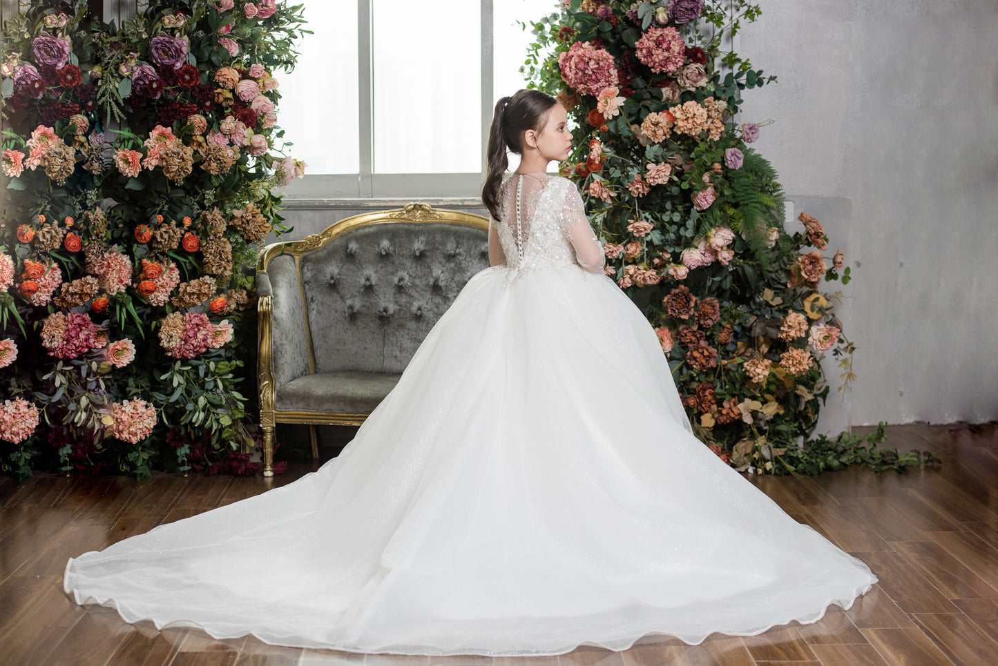 Woman in a white wedding dress standing in front of floral decorations and a chair.
