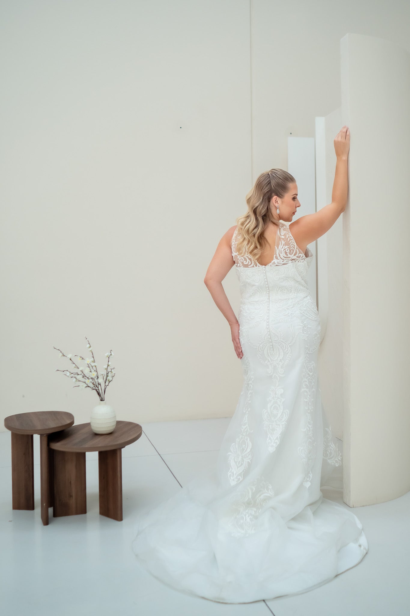 Woman in a white lace wedding dress standing in a minimalistic room with wooden tables and a vase.