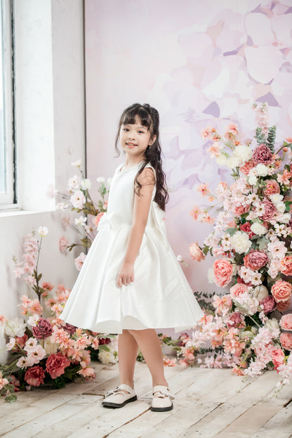 Young girl in a white dress standing in front of floral decorations.