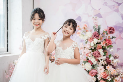 Two young girls in white dresses standing in front of a floral arrangement.