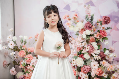 Young girl in a white dress holding a flower in front of a floral backdrop