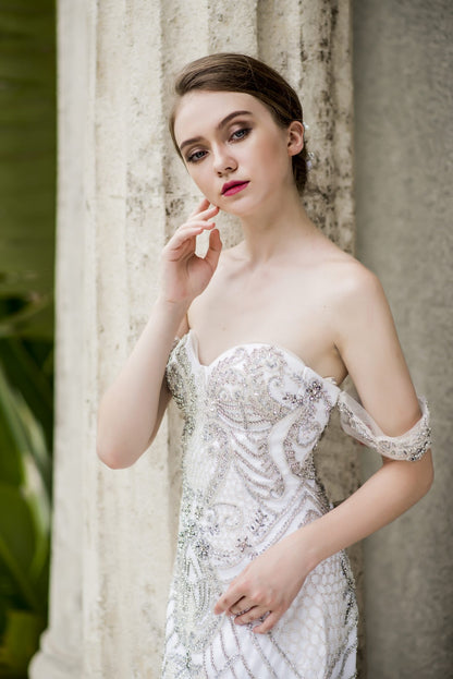 Woman in a white strapless dress with floral embellishments standing against a textured wall.