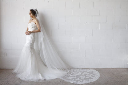 Woman in a white wedding dress with a long veil standing against a white brick wall.