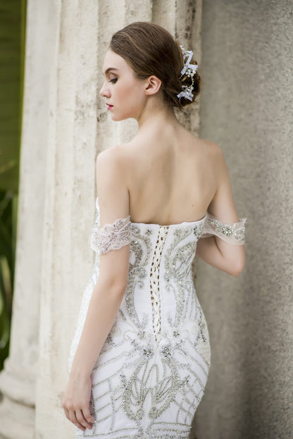 Woman in a white, off-shoulder wedding dress with intricate detailing standing against a stone wall.