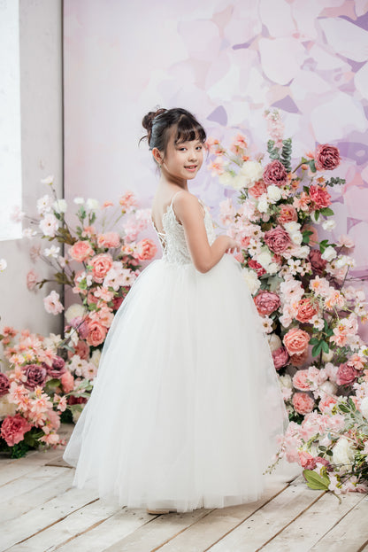 Young girl in a white dress standing in front of a floral arrangement with a pastel-colored background.