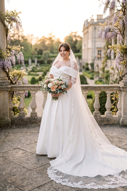 A bride in a white off-the-shoulder wedding dress with a long lace-trimmed veil holds a floral bouquet on a stone terrace with a garden and historic building in the background.