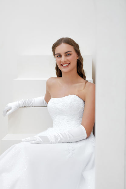 Woman in a white wedding dress and gloves posing against a white wall.