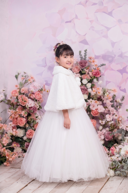 A young girl wearing a white tulle dress with a fluffy white cape stands in front of a pink floral backdrop.