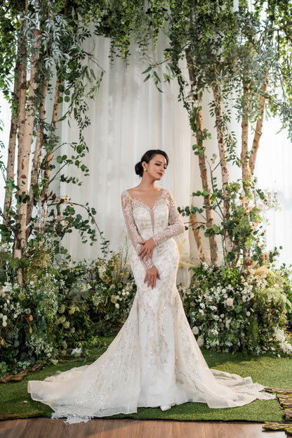 Woman in a white lace wedding dress standing in a forest with birch trees and greenery.