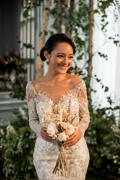 Bride in a lace wedding dress holding a bouquet of flowers, surrounded by greenery.