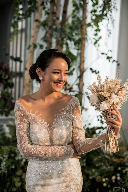 Woman in a wedding dress holding a bouquet of flowers, standing outdoors with greenery in the background.