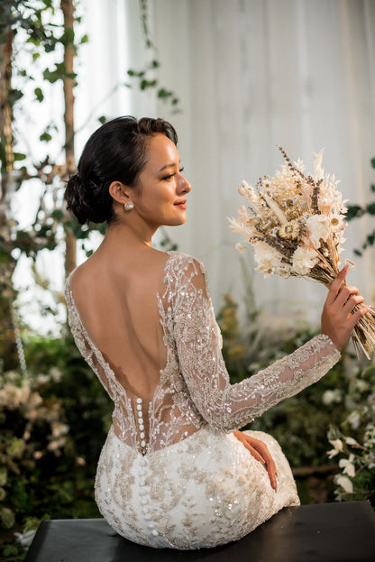 Bride in a white wedding dress with a low back, holding a bouquet of flowers, sitting outdoors.