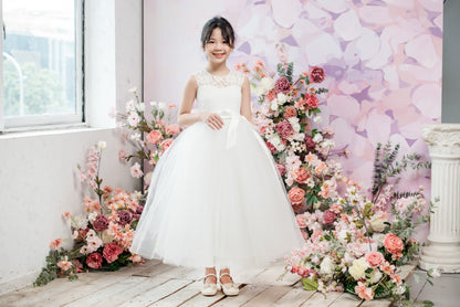 Young girl in a white dress standing in front of floral decorations and a pastel-colored wall.