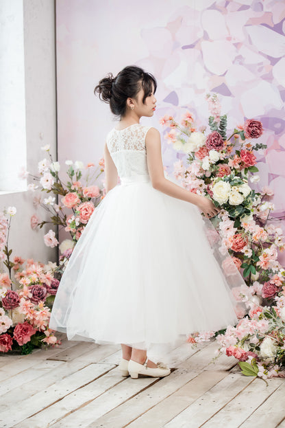 Woman in a white dress standing in front of a floral arch with a light purple wall.