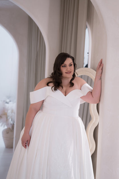Woman in a white off-shoulder dress standing in an elegant interior setting.