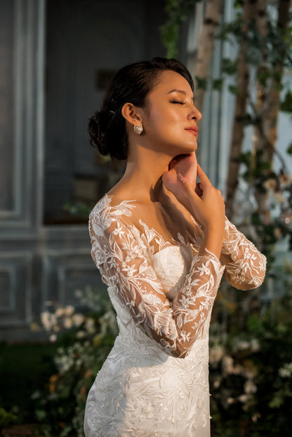 Woman in a white lace dress standing outdoors with sunlight filtering through.