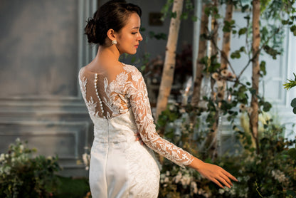 Woman in a white lace wedding dress standing outdoors with greenery and a building in the background