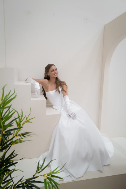 Woman in a white wedding dress sitting on a white bench with greenery in the foreground.