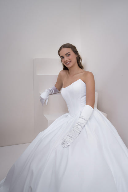 Woman in a white wedding dress with gloves sitting on a chair against a plain background
