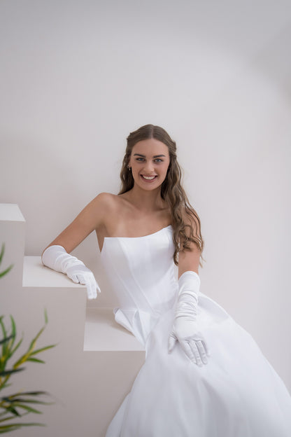 Woman in a white wedding dress with gloves posing against a plain background
