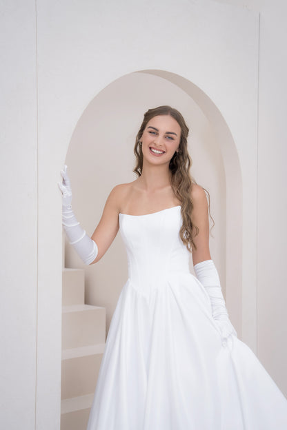 Woman in a white wedding dress with long gloves standing in a softly lit room.