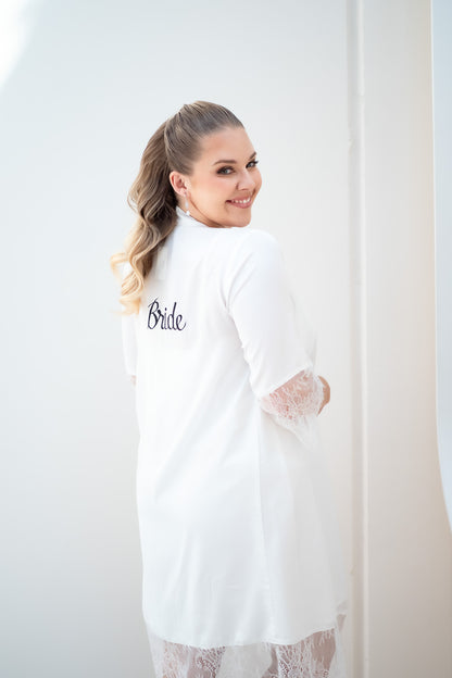 Woman wearing a white robe with 'Bride' embroidered on it, standing against a white curtain