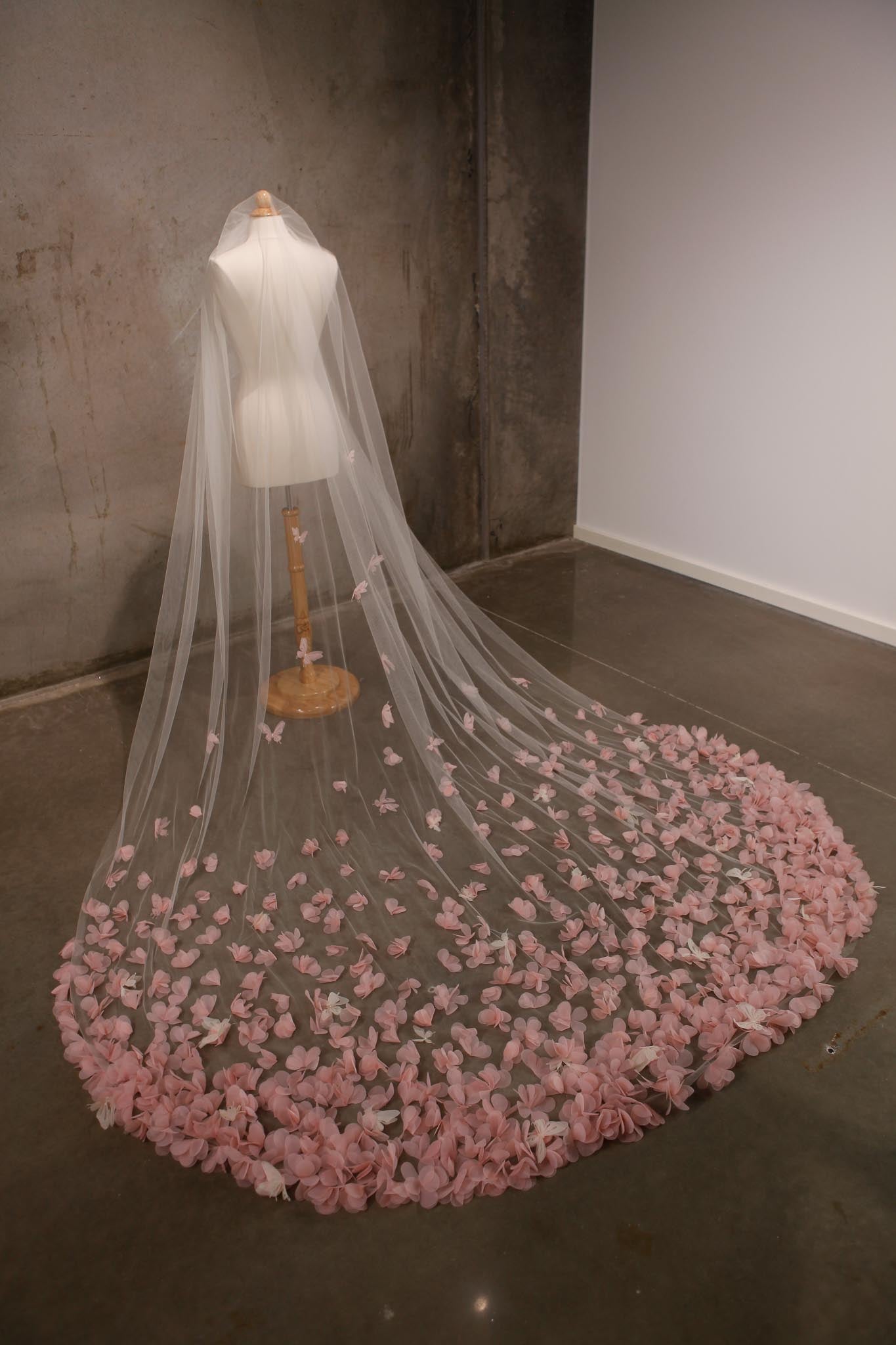 Long white veil with pink flower petals on a mannequin against a plain wall.