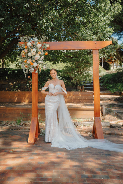 Woman in a wedding dress standing under a wooden arch with floral decorations in an outdoor setting