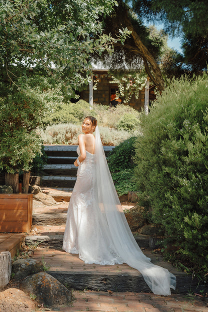 Woman in a wedding dress standing in a garden setting with greenery and a house in the background
