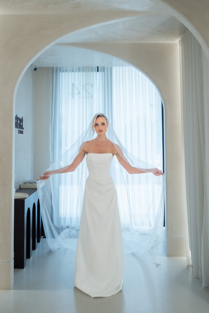 Woman in a white wedding dress standing in a room with large windows and white curtains.