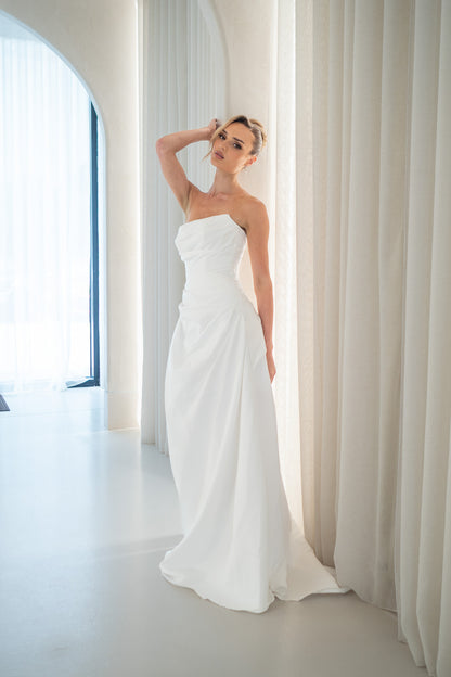 Woman in a strapless white wedding dress standing in a bright room with large windows.