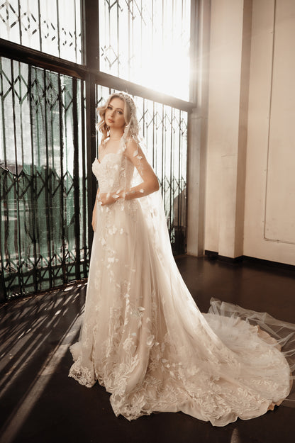 Woman in a white lace wedding dress standing in a room with large windows.