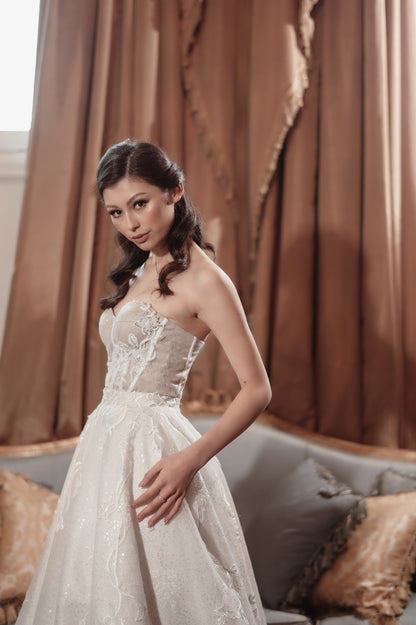 Woman in a white lace dress standing in a room with brown curtains and a sofa.