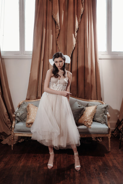 Woman in a white dress standing in a room with brown curtains and a wooden floor.