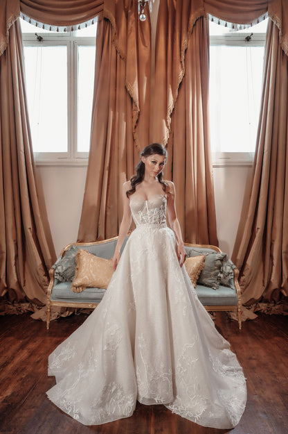 Woman in a white lace wedding dress standing in a room with large brown curtains and a bench.