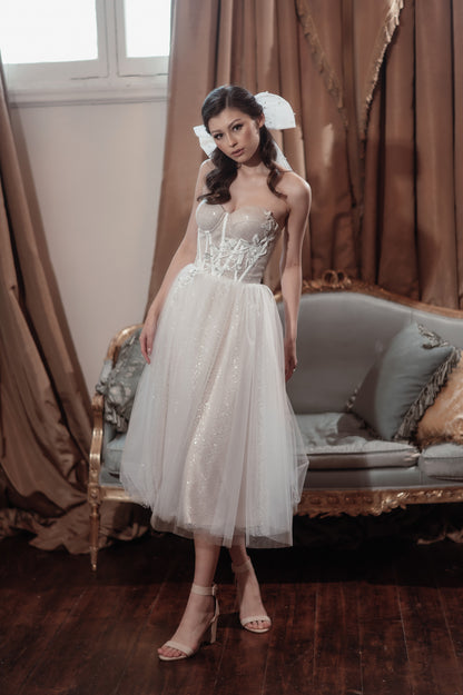 Woman in a white dress standing in a room with curtains and a sofa.