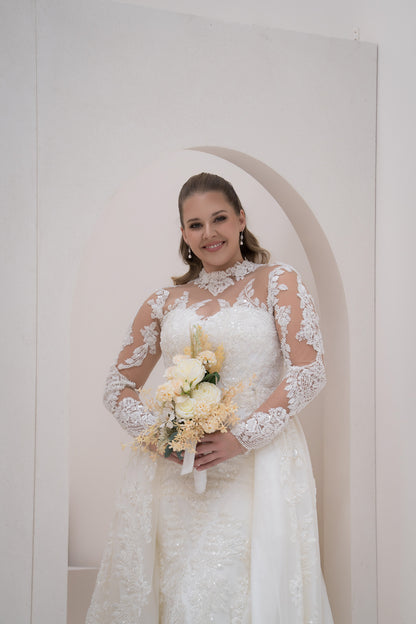 Woman in a white lace wedding dress holding a bouquet against a plain background