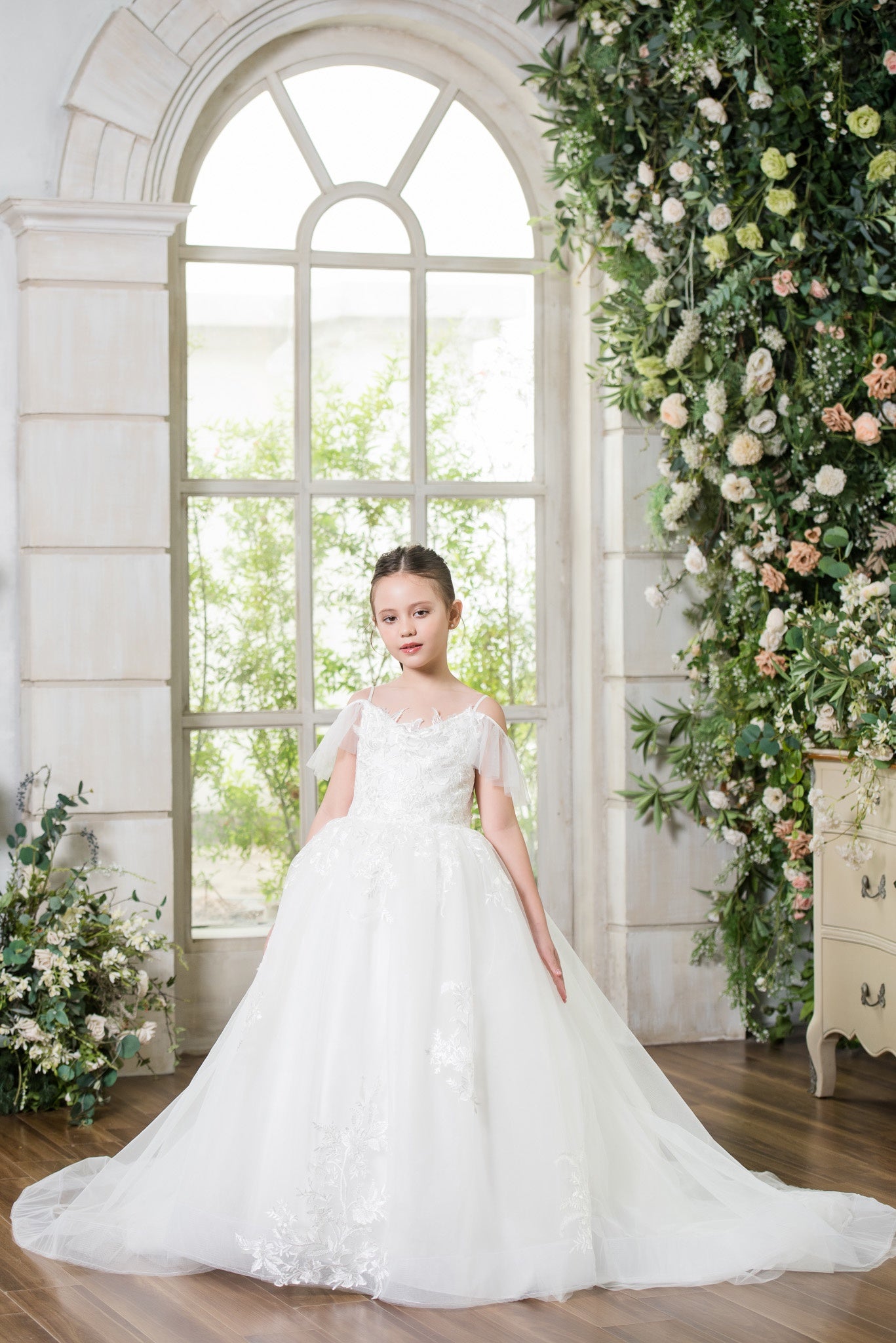 Young girl in a white dress standing in a decorated room with flowers and a large window.