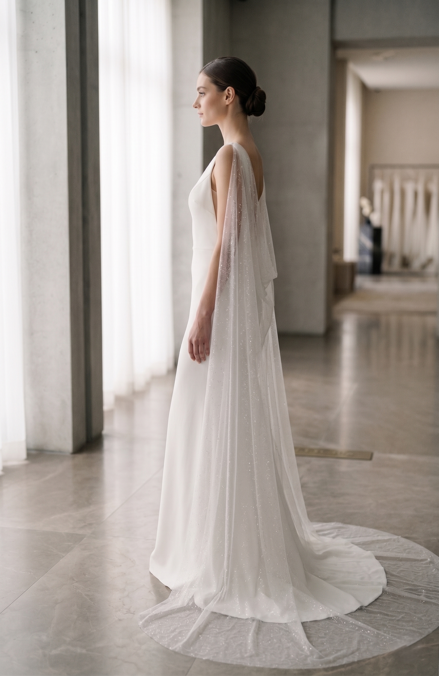 Woman in a white wedding dress standing in a softly lit room with large windows.