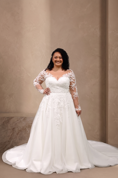 A woman modeling a white wedding gown with long sheer lace sleeves, a fitted lace bodice, and a full flowing skirt with a train, posed against a warm beige textured wall.