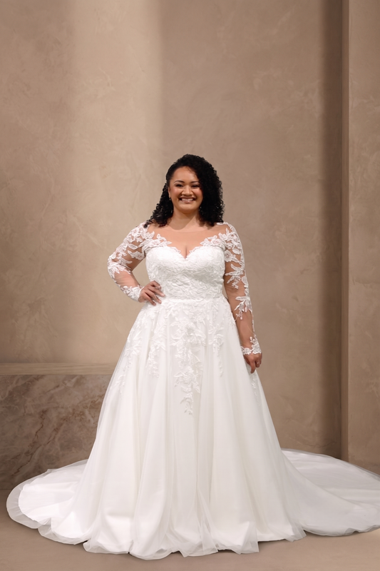 A woman modeling a white wedding gown with long sheer lace sleeves, a fitted lace bodice, and a full flowing skirt with a train, posed against a warm beige textured wall.