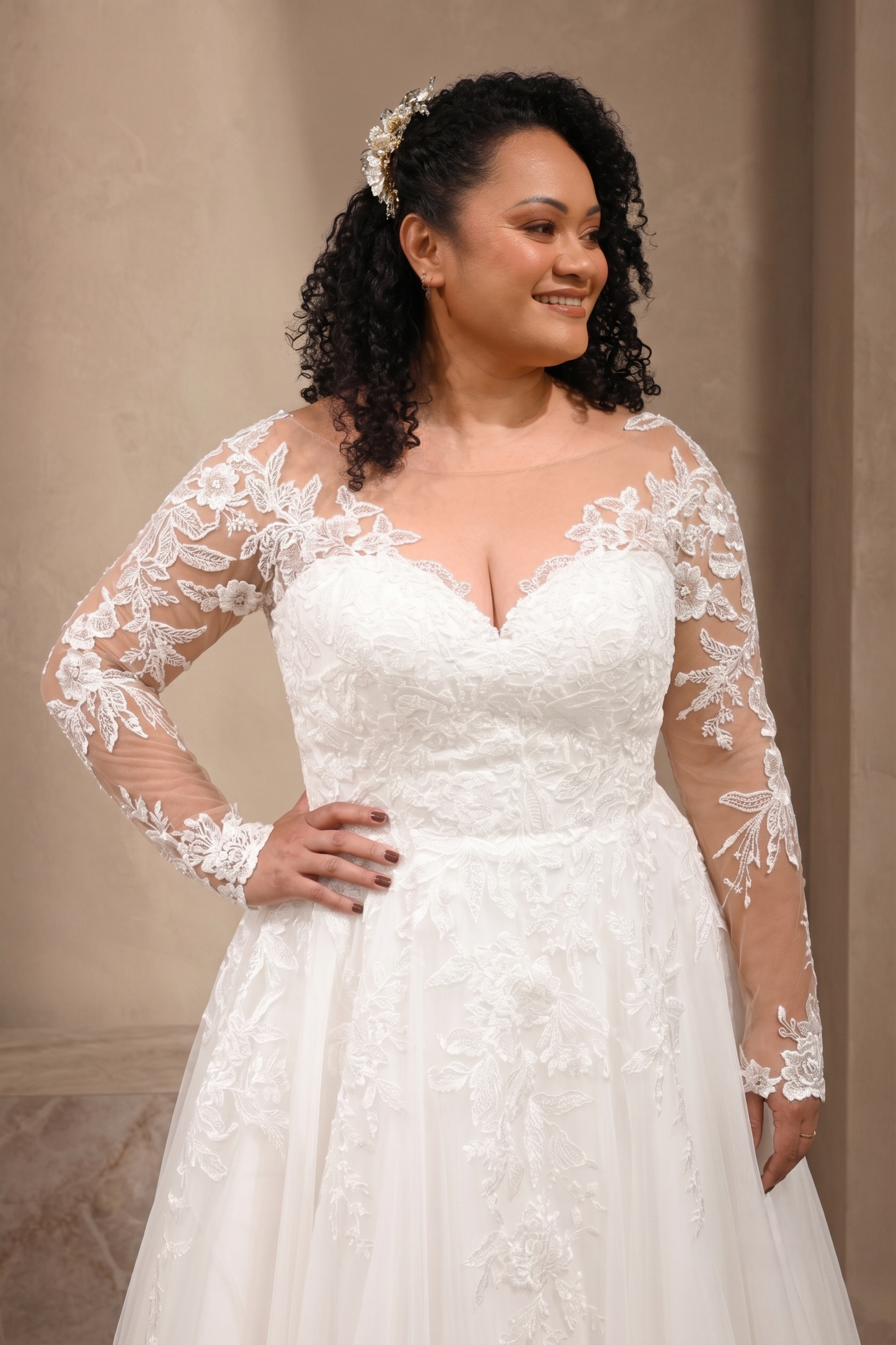 A woman modeling a white lace wedding dress with long sheer sleeves, a sweetheart neckline, and a full skirt against a warm beige indoor backdrop.