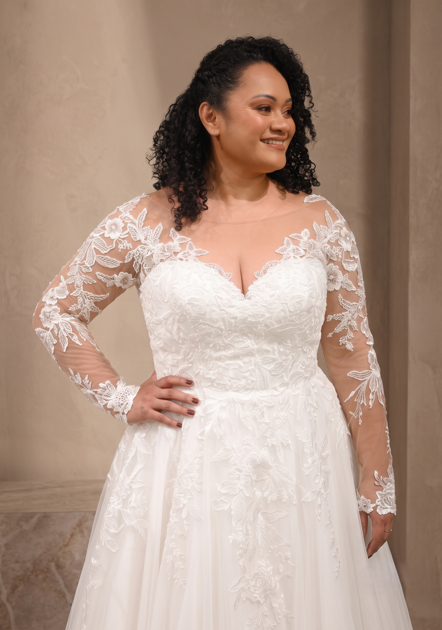 A woman modeling a white lace wedding gown with long sheer lace sleeves, a sweetheart neckline, and a full skirt against a beige indoor backdrop.