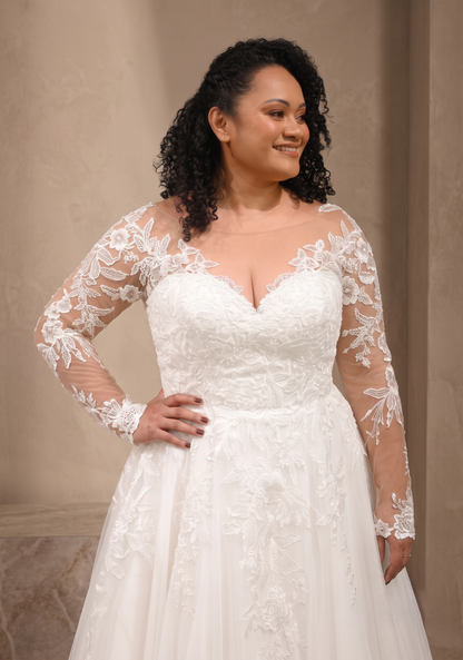 A woman modeling a white lace wedding gown with long sheer lace sleeves, a sweetheart neckline, and a full skirt against a beige indoor backdrop.
