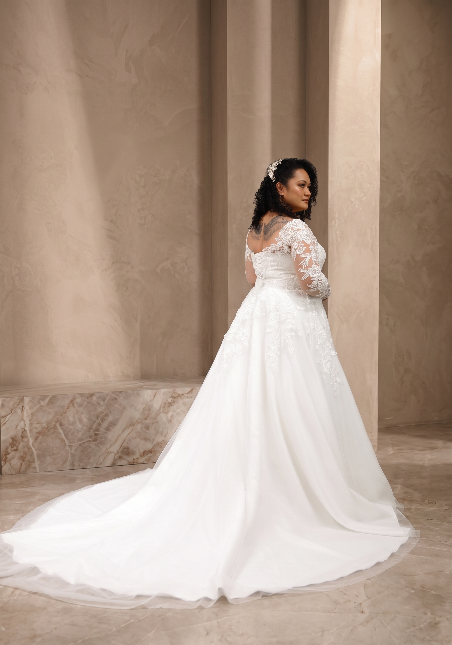 A woman wearing a white wedding gown with lace long sleeves and a full tulle skirt, standing in a softly lit interior with beige stone walls and columns.