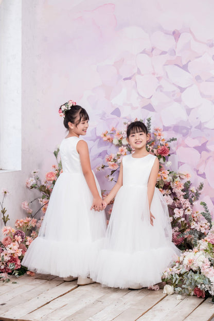 Two young girls in white dresses standing in front of a floral backdrop with a soft pink and purple color scheme.