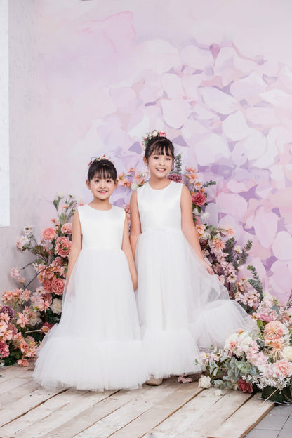 Two young girls in white dresses standing in front of a floral arrangement with a decorative background.