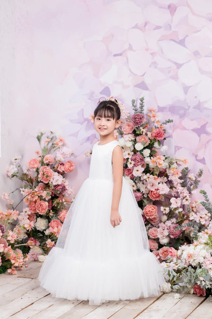Young girl in a white dress standing in front of a floral backdrop with pink and purple flowers.