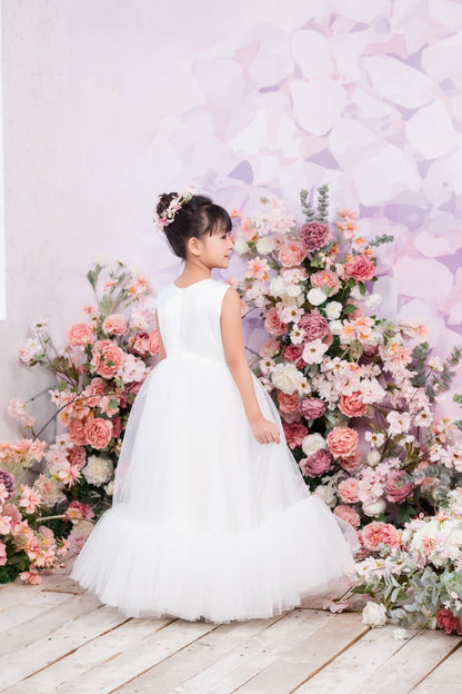 Young girl in a white dress standing in front of a floral arrangement with a light purple background